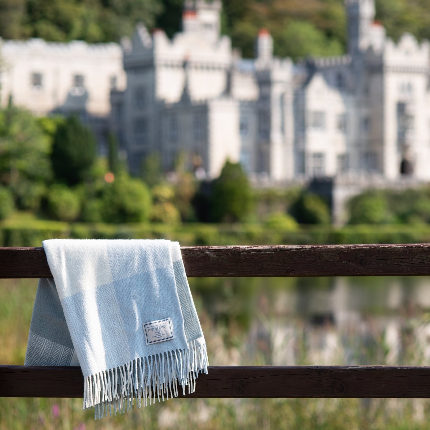 A blue and white cashmere throw draped over a fence with a scenic view of Kylemore Abbey in the background.